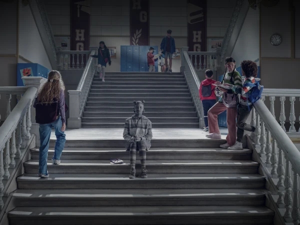 A group of teenagers are standing and walking on the stairs of a high school, with banners in the background and neutral lighting.